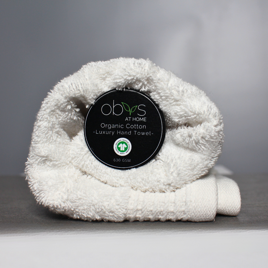 A neatly folded organic cotton towel in a soft, natural shade, with a smooth, plush texture. The towel lies on a wooden surface, radiating a sense of warmth and luxury. Nearby, a hint of greenery adds a touch of nature, highlighting the towel’s eco-friendly, sustainable origins, perfect for a relaxing, spa-like self-care experience