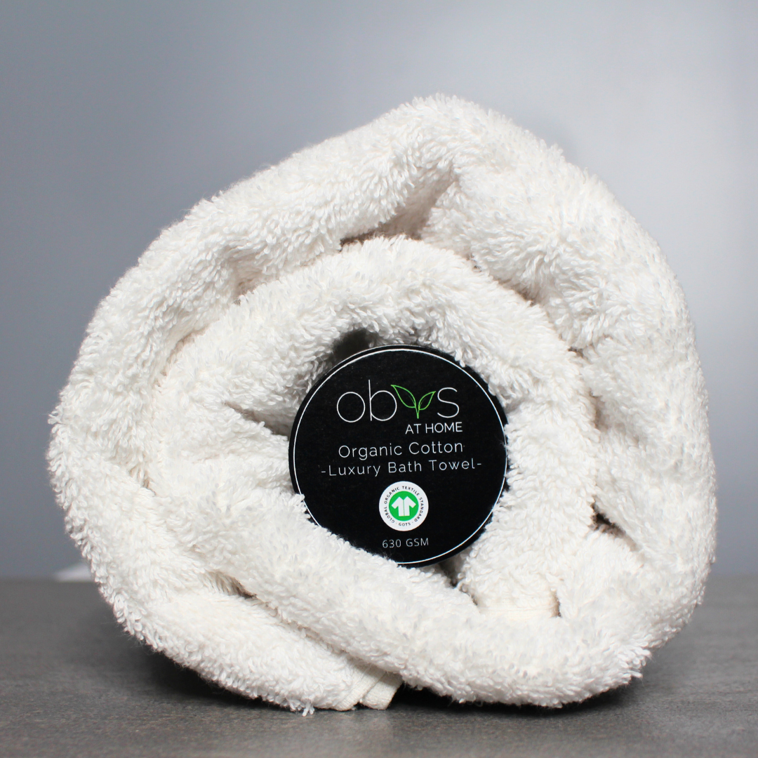 A neatly folded organic cotton towel in a soft, natural shade, with a smooth, plush texture. The towel lies on a wooden surface, radiating a sense of warmth and luxury. Nearby, a hint of greenery adds a touch of nature, highlighting the towel’s eco-friendly, sustainable origins, perfect for a relaxing, spa-like self-care experience