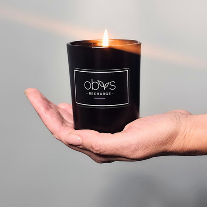 A matte black candle jar filled with organic essential oils, burning with a soft, warm flame. The minimalist design exudes elegance, while the gentle aroma from natural essential oils creates a calming, eco-friendly atmosphere. The candle sits on a wooden surface, with subtle shadows enhancing its luxurious, earthy vibe, perfect for relaxation or meditation