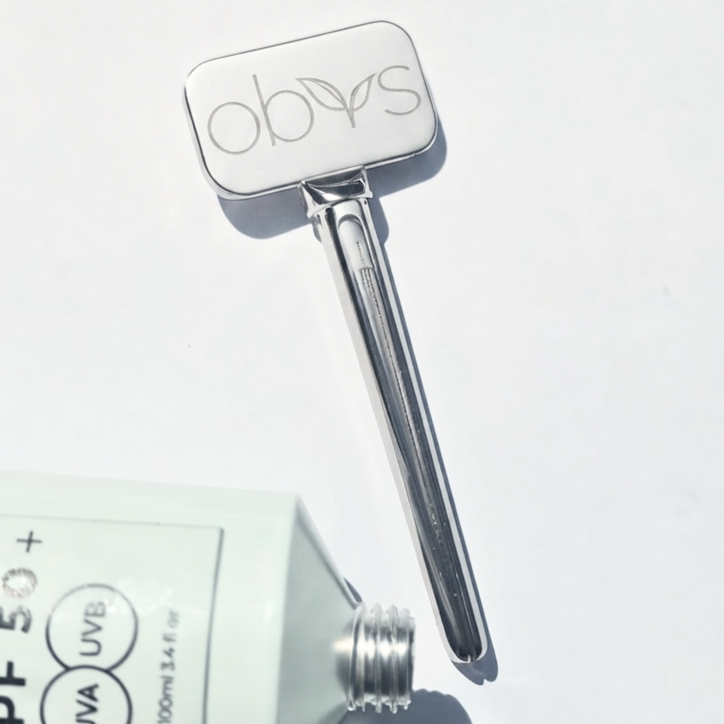 A silver metal beauty tool with a rounded rectangular head engraved with the brand name 'obys' and a long tapered handle, shown on a white background next to a partially visible sunscreen tube.