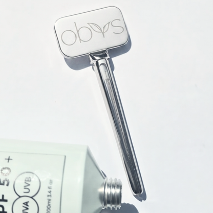 A silver metal beauty tool with a rounded rectangular head engraved with the brand name 'obys' and a long tapered handle, shown on a white background next to a partially visible sunscreen tube.