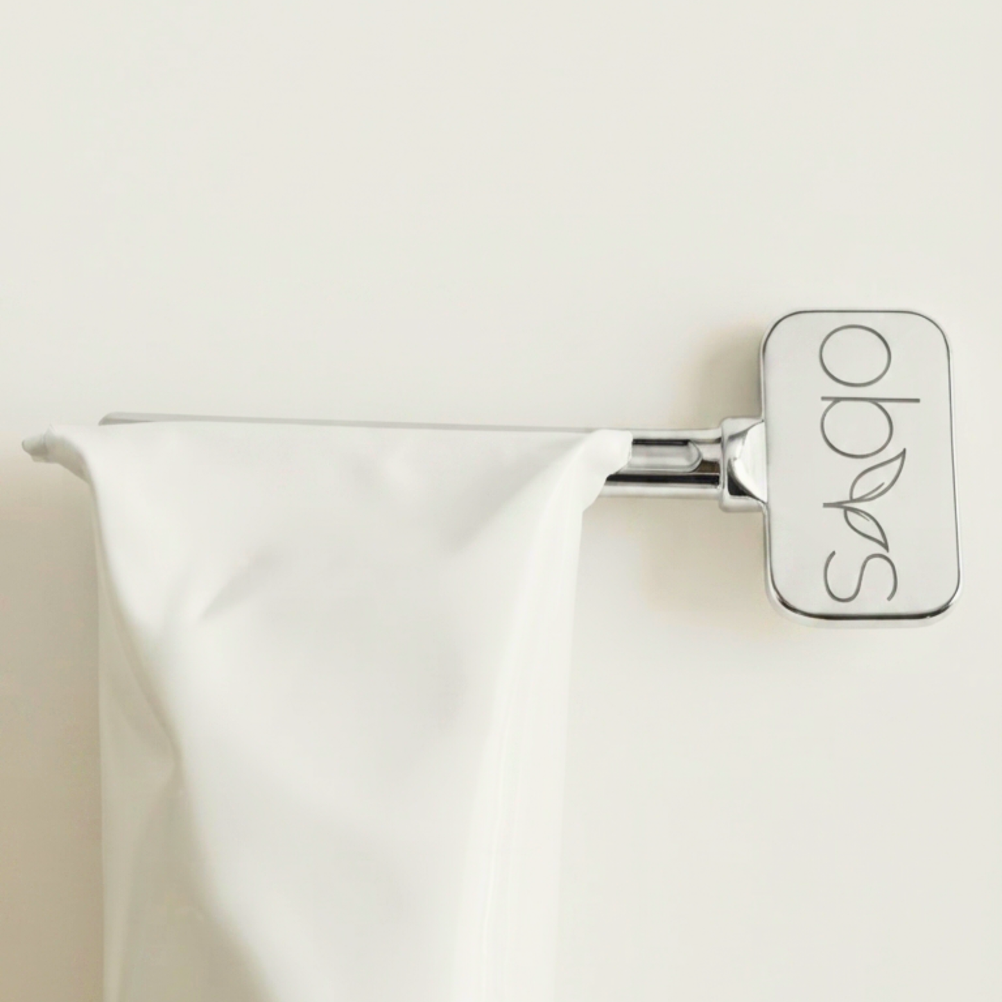 A polished chrome towel bar with a rectangular end cap engraved with the brand name 'soho' and leaf motifs, holding a white towel against a plain off-white wall.
