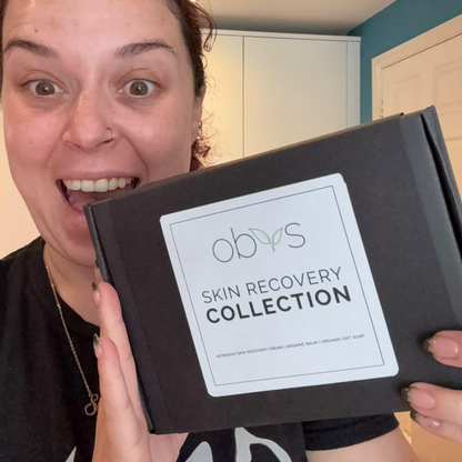 Dani Harmer Tracy Beaker holding a black box labeled 'obys Skin Recovery Collection' in a home setting with white cabinets and a teal wall in the background.