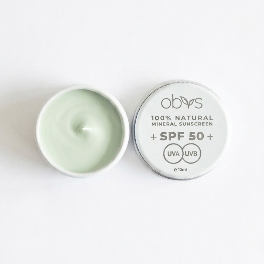 An open round metal tin of Obys 100% Natural Mineral Sunscreen SPF 50+ with a pale mint green cream inside, shown next to its silver lid on a clean white background.