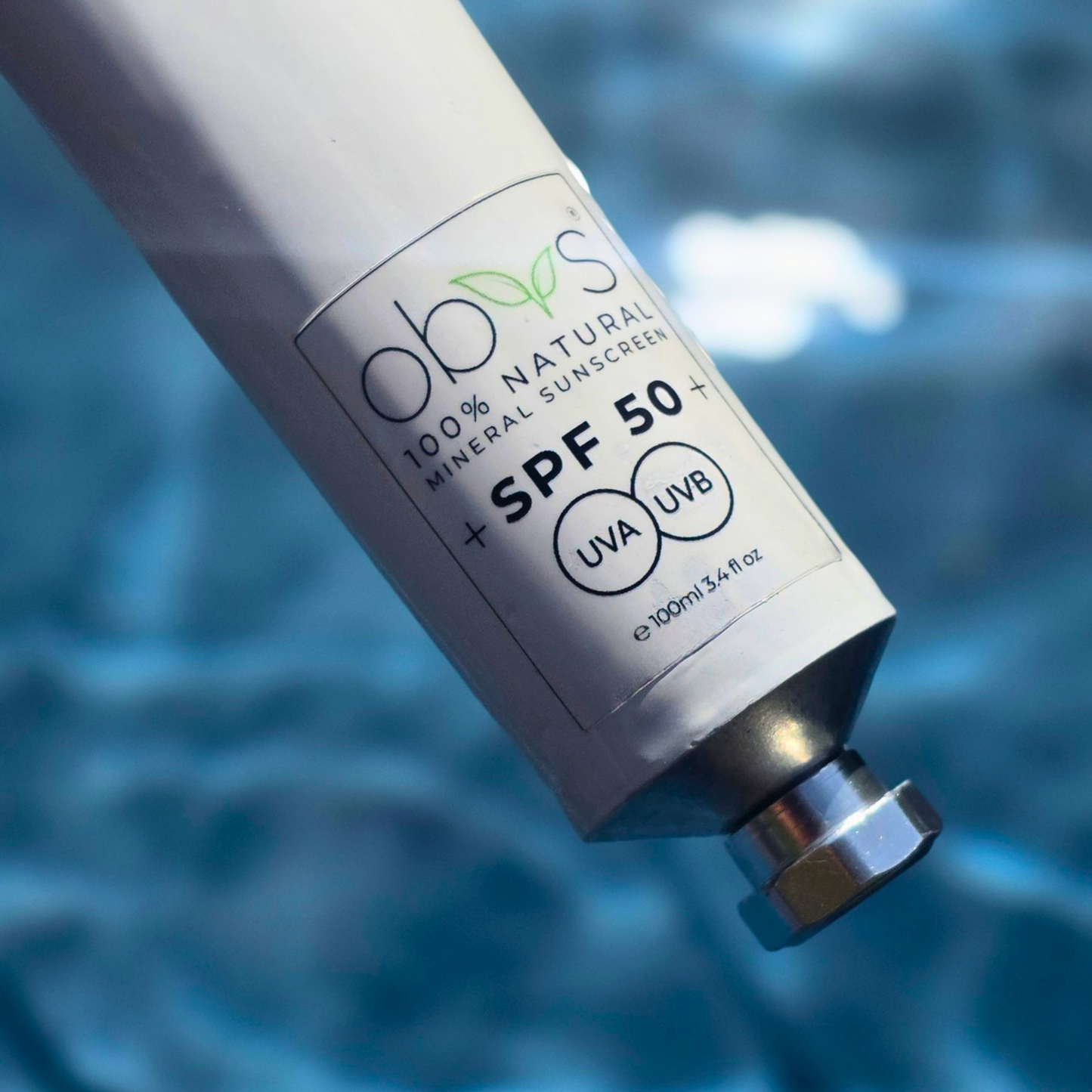 A close-up of a white sunscreen tube labeled 'obios 100% Natural Mineral Sunscreen SPF 50+' with a silver cap, set against a blurred blue water background.