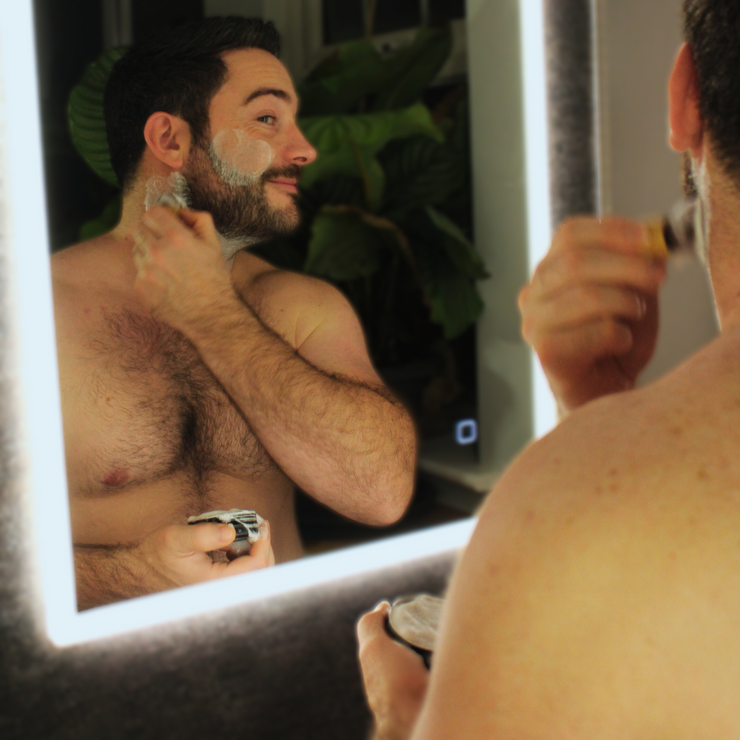 Man shaving his beard in front of a mirror with a lighted border Obvs Skincare New Shaving Soap Travel Kit with peppermint and charcoal shaving soap in travel tin and mini shaving brush