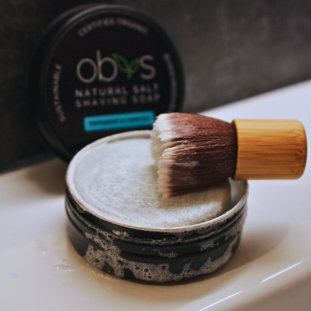 Shaving brush and soap on a white surface with 'obvs' branding.Obvs Skincare New Shaving Soap Travel Kit with peppermint and charcoal shaving soap in travel tin and mini shaving brush