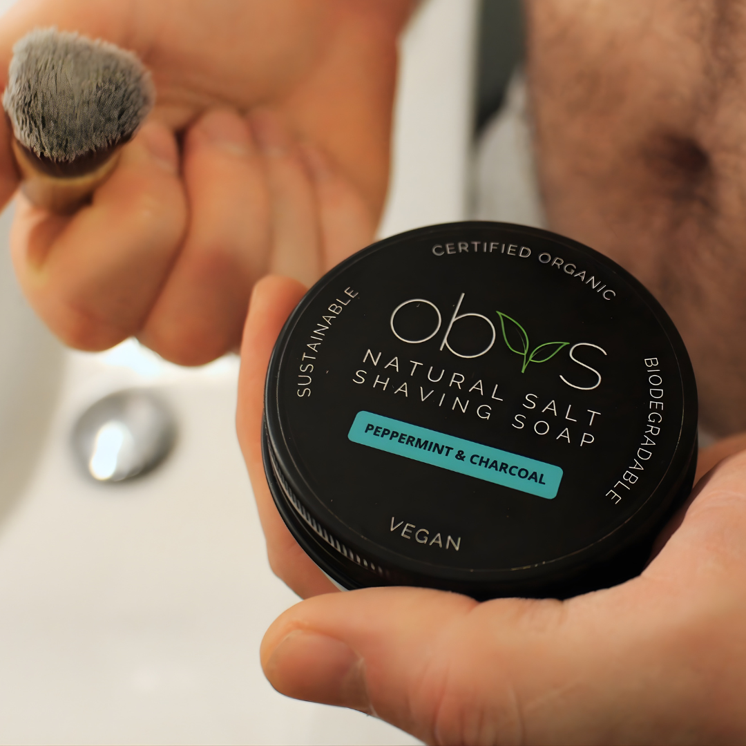 Person holding a container of Obv's natural salt shaving soap with a brush.  Obvs Skincare New Shaving Soap Travel Kit with peppermint and charcoal shaving soap in travel tin and mini shaving brush
