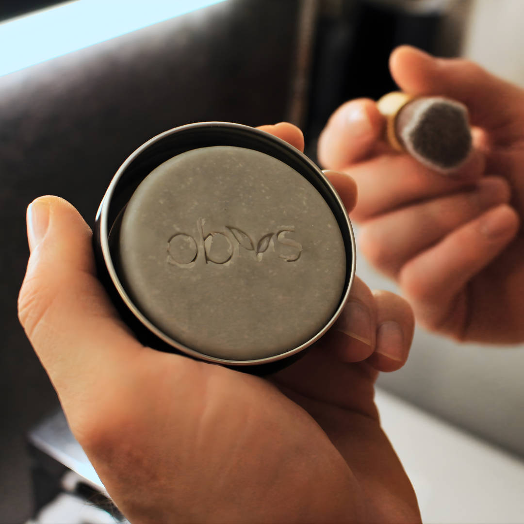 Hand holding a container of soap with 'obvs' branding. Obvs Skincare New Shaving Soap Travel Kit with peppermint and charcoal shaving soap in travel tin and mini shaving brush